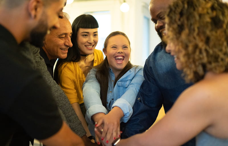 A woman with down syndrome huddles up with a group of people and put their hands in the middle of the group circle.