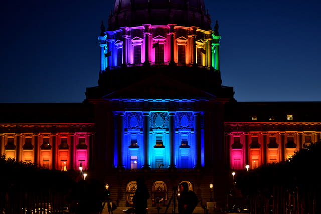 a picture of a city hall building, lighted with rainbow colored lights in honor of gay pride