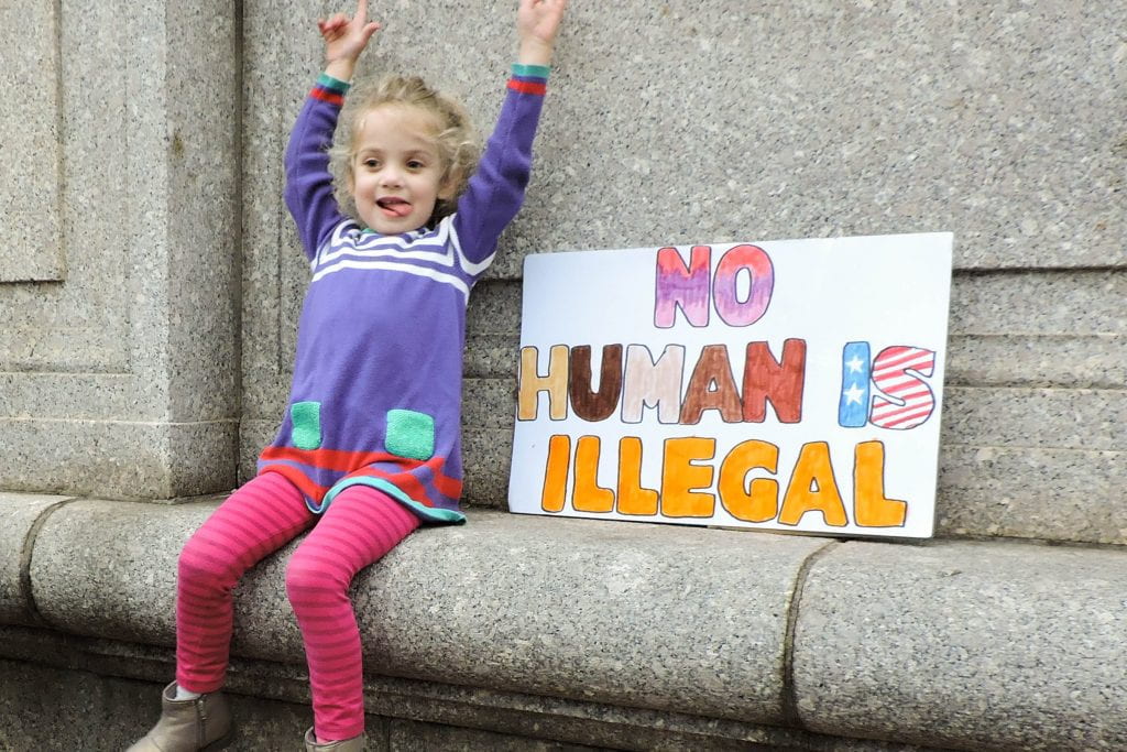 A young girl with her hands in the air and tongue stuck out in a silly expression sits beside a sign reading, "No human being is illegal!'