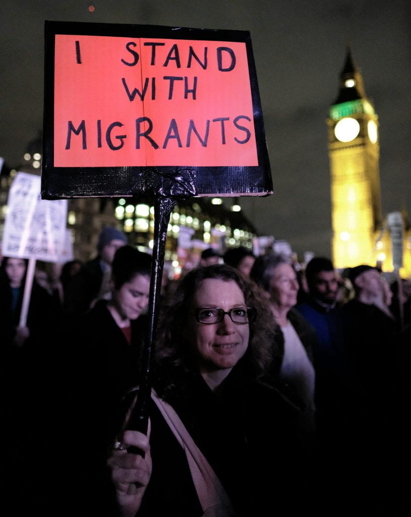 A woman at a protest in London holds a sign saying, "I stand with migrants."