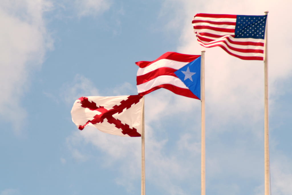 The American flag, Puerto Rican flag, and Spanish flag are shown flying in front of a blue sky.