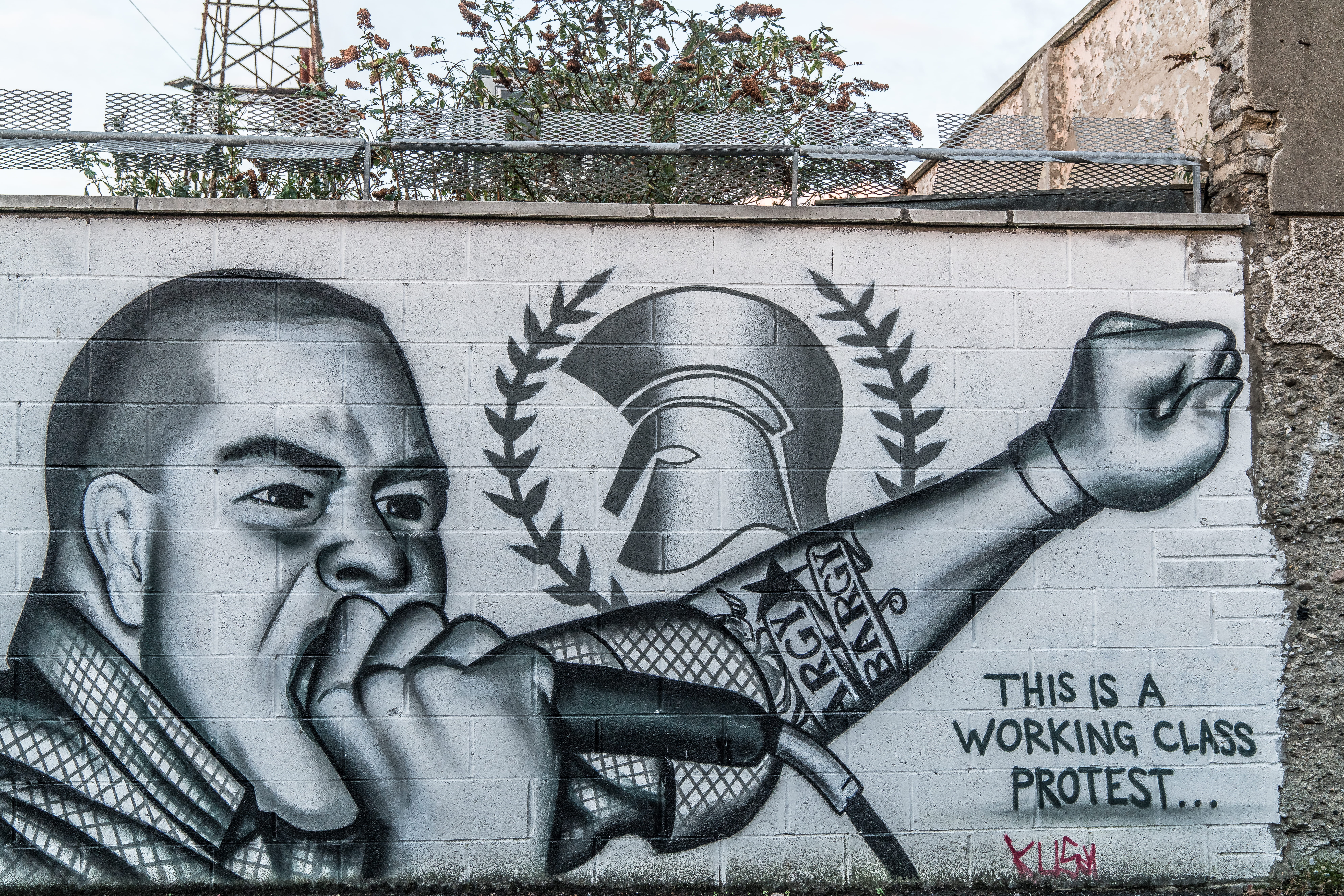 White wall with black and white graffiti of a man holding a microphone with fist in the air. Text at the bottom of the graffiti says, "This is a working class protest..."