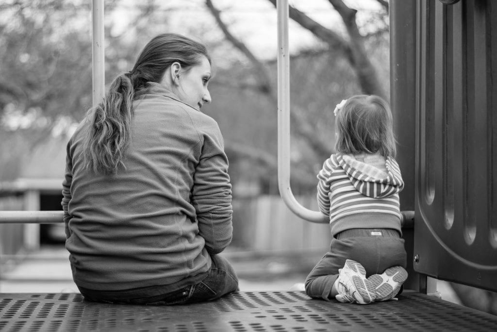 A woman sits on a playground next to her young daughter. 