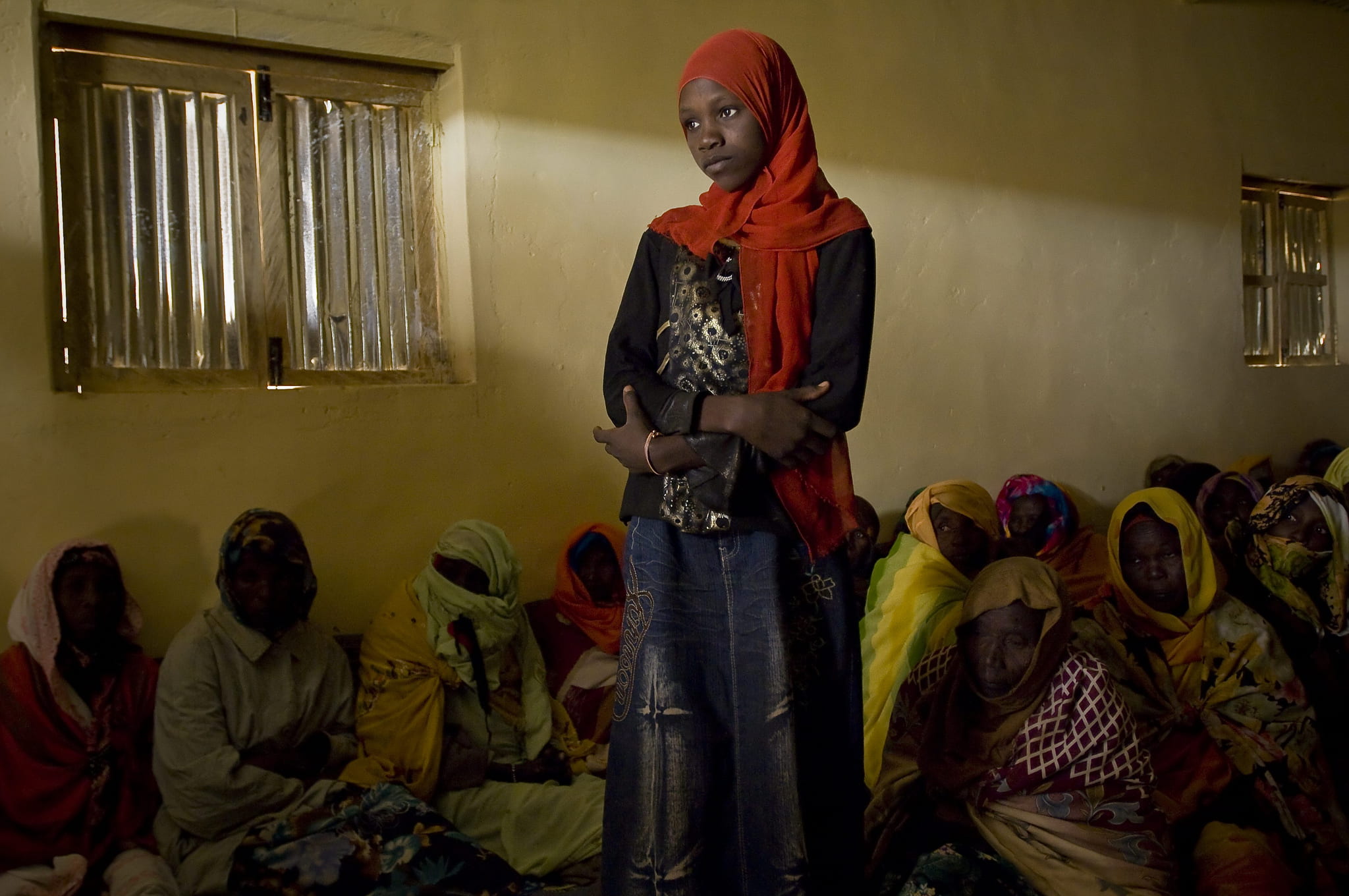 A refugee woman in a bright red hijab stands in a dark room with other women seated on the floor behind her.