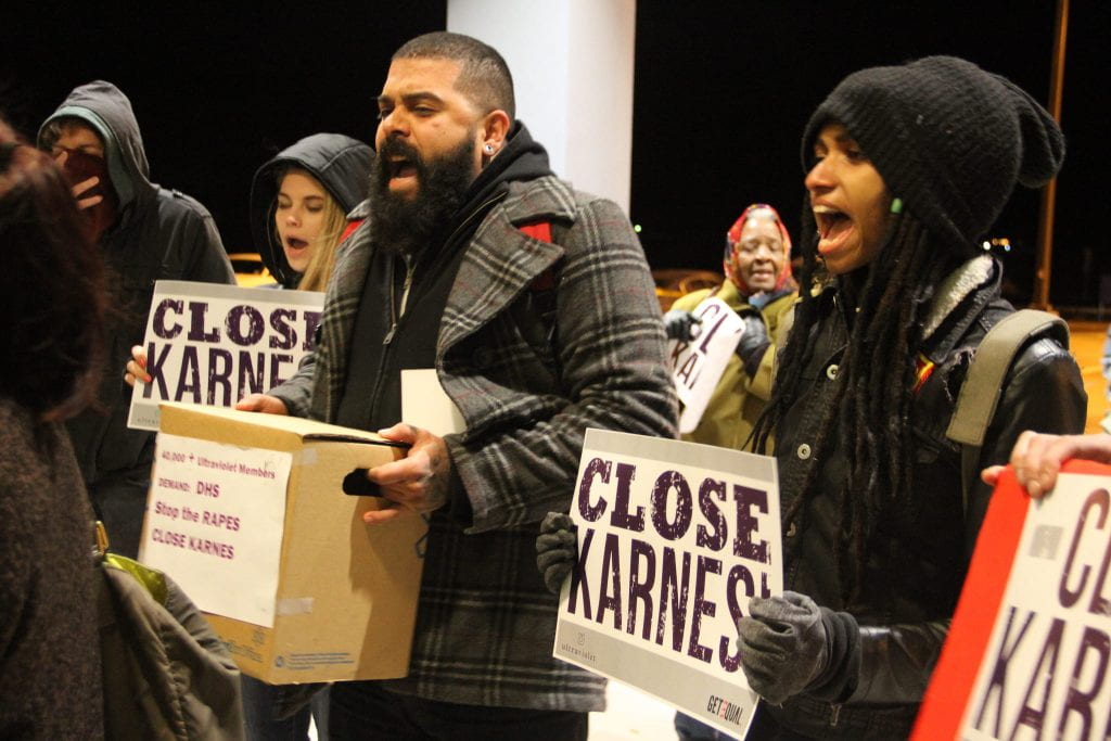 A crowd of people appear to be yelling as they hold signs that say "Close Karnes."