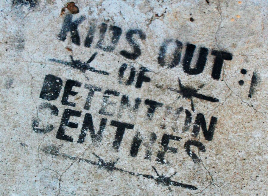 "Kids Out of Detention Centers" is stamped onto concrete in black ink with barbed wires surrounding the text. 