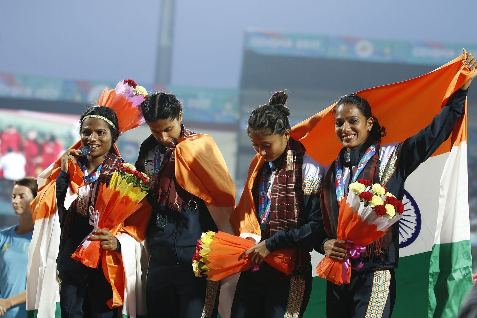 Dutee Chand bites her medal on the left while standing with her three teammates.