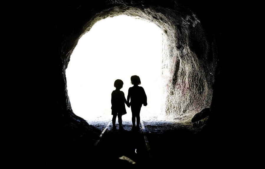 Two children stand in a circular entrance to a tunnel holding hands. 