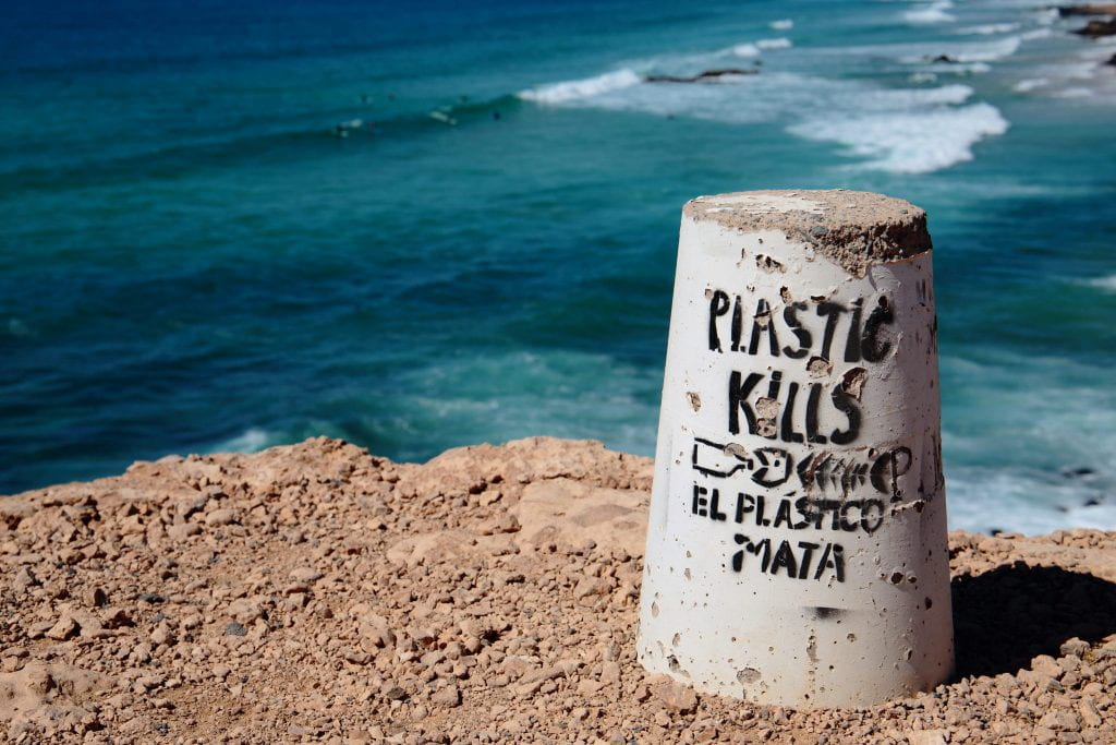 A concrete cylinder sits on the right of the shot, in front of ocean waves. The cylinder has "plastic kills! El plastico mata !"
