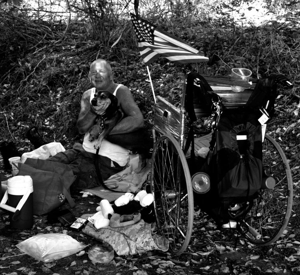 A homeless man with disabilities sits on the ground with his dog. His wheelchair is next to them, along with all his items and an American flag. 