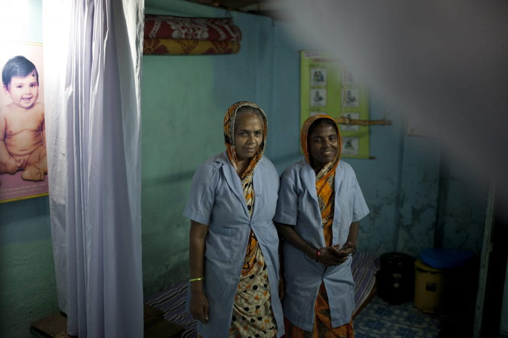 Two midwives stand next to a curtain in a dim room with photos of babies on the walls.