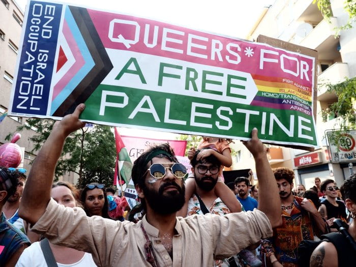 A man holds a rainbow Pride protest sign with phrases including “End Israeli occupation”, “Queers for a free Palestine”, and “Fight against: racism, Islamophobia, homo/transphobia, antisemitism, apartheid”