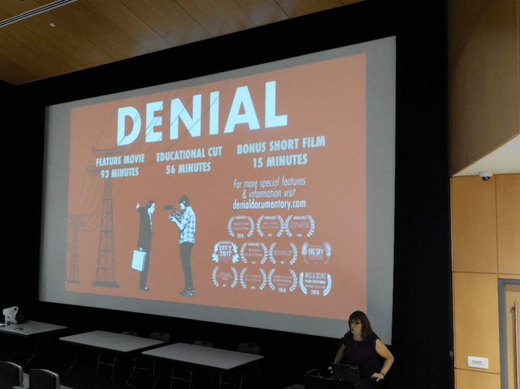 Christine Hallquist, in front of a screen showing her film "Denial" talking to the audience.