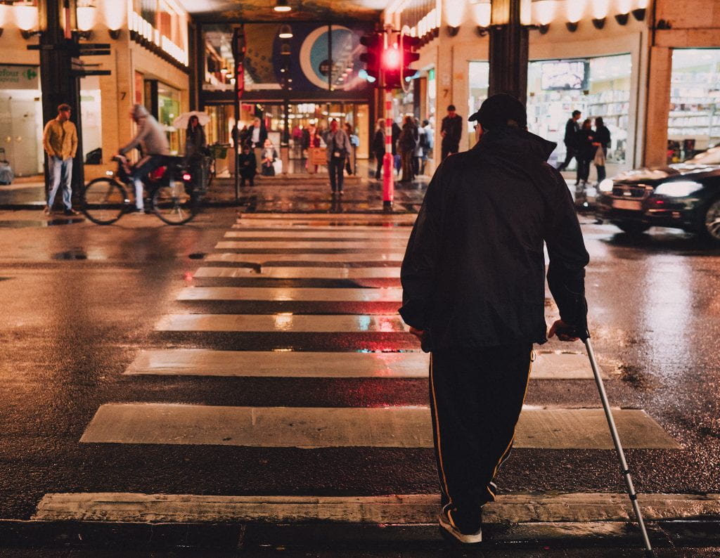 A man in dark clothing crosses a city street with the aid of a walking stick / mobility device of some sort. 