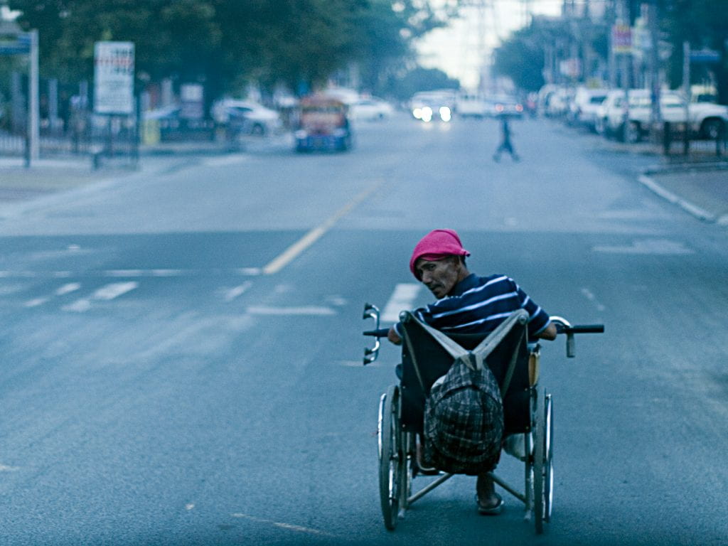 A man in a wheelchair looks back as he wheels down the street. He is wearing a bright pink hat and has a backpack hanging off the handles of his chair. 