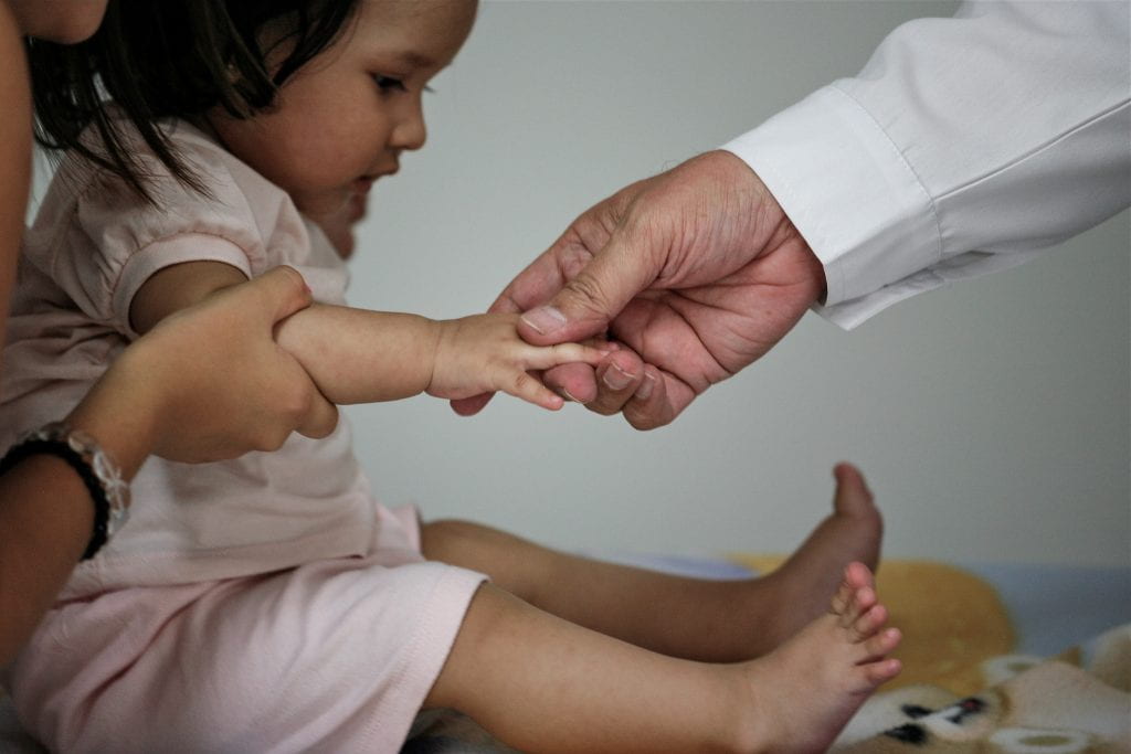 A young child in a pink dress has her fingers held by a white-sleeved hand for an examination.