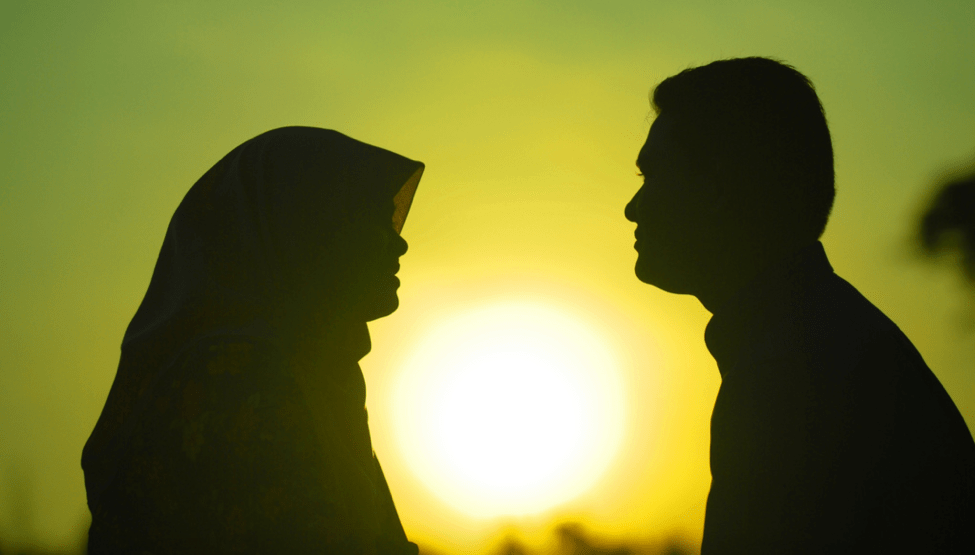 The silhouettes of a man and a hijabi woman face each other with a sunset behind them