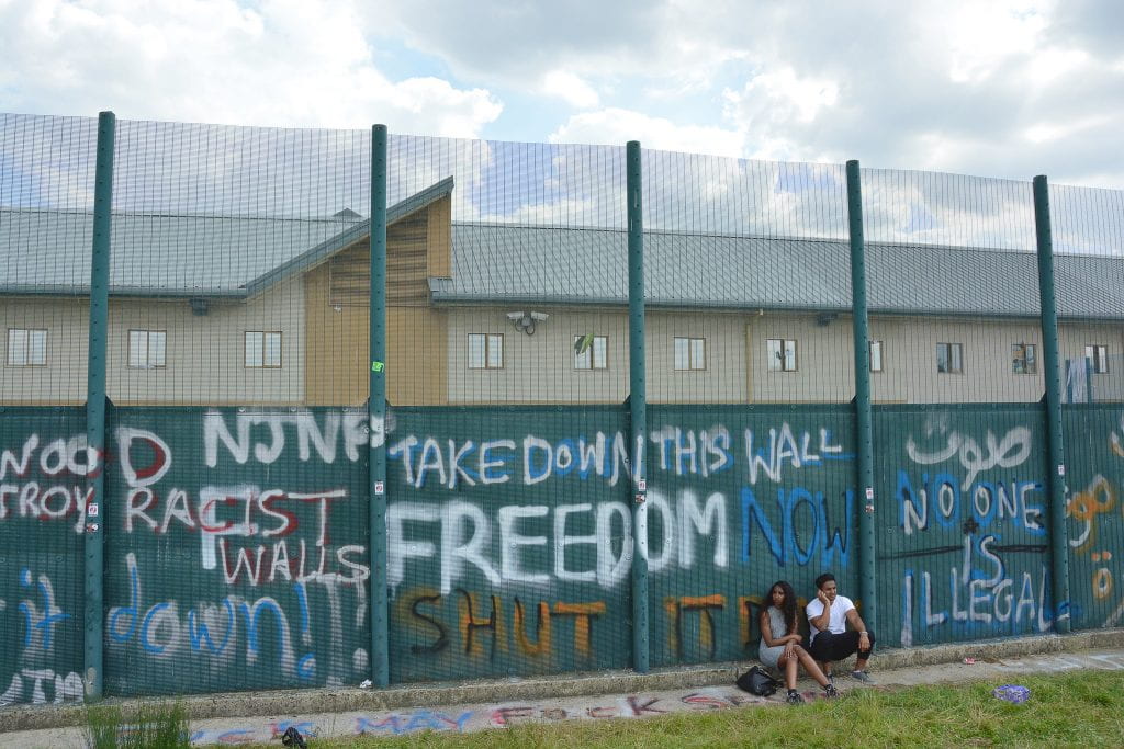 A couple sits next to a large wall with graffiti saying "Freedom," and "Take down this wall!"