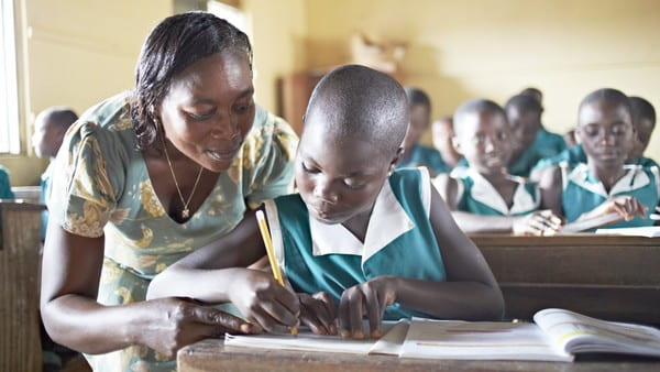 A teacher looks on as a young African girl does her school work.