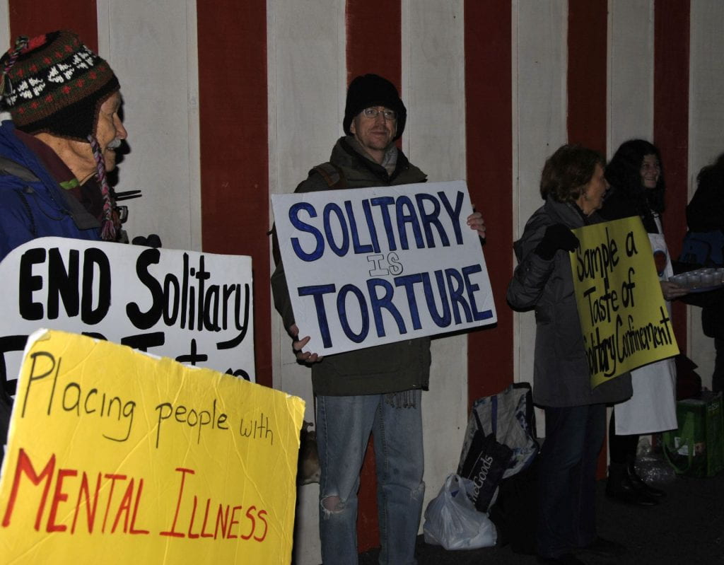 Image of protesters of solitary confinement holding signs connecting solitary confinement to torture and mental illness.
