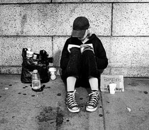A homeless student, sitting on the sidewalk against a wall, reading a book. The student has a small bag of items beside him and a sign that says, "Homeless."