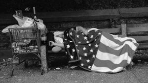 A homeless man sleeps under an American flag blanket on a park bench in New York City.