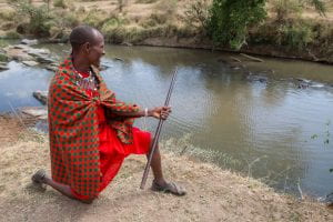 A Maasai man in traditional red clothing overlooks the Sekenani River. Nearby vegetation reflects off the water's rippled surface.