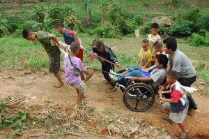a group of young children help push a man in a wheelchair up a dirt road