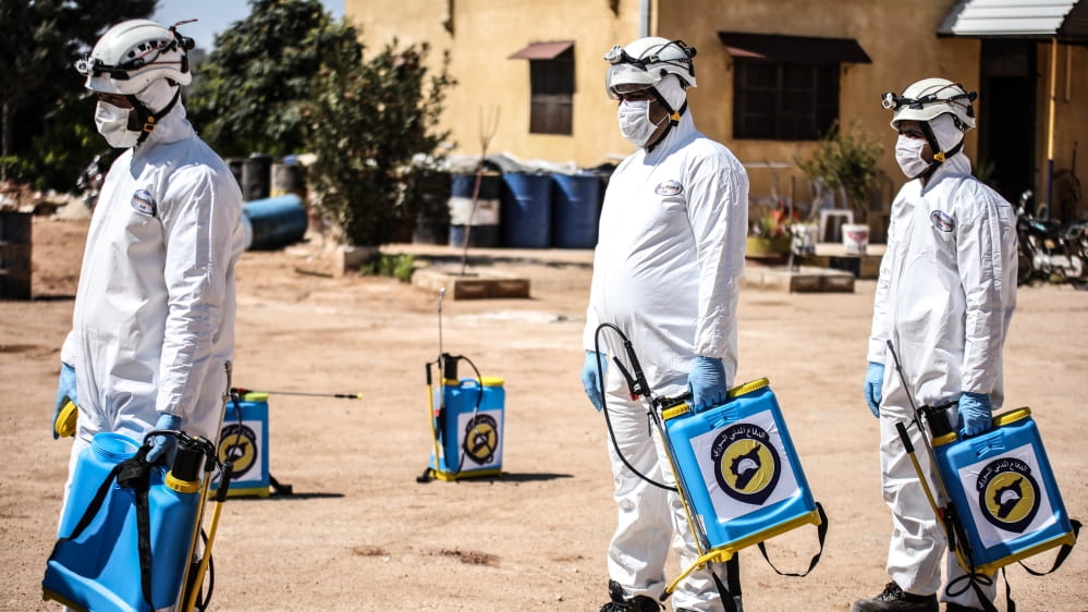 A photo of 3 medical professionals in masks and white suits carrying testing machines in war-torn Syria