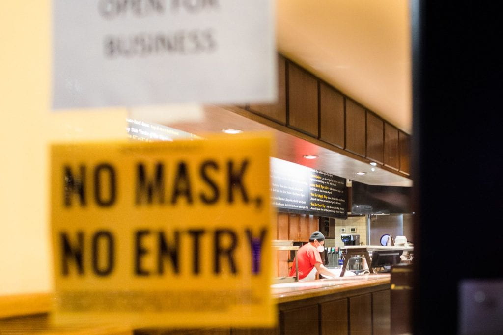 Person with a protective mask preparing food with a front door sign that reads "No Mask, No Entry".