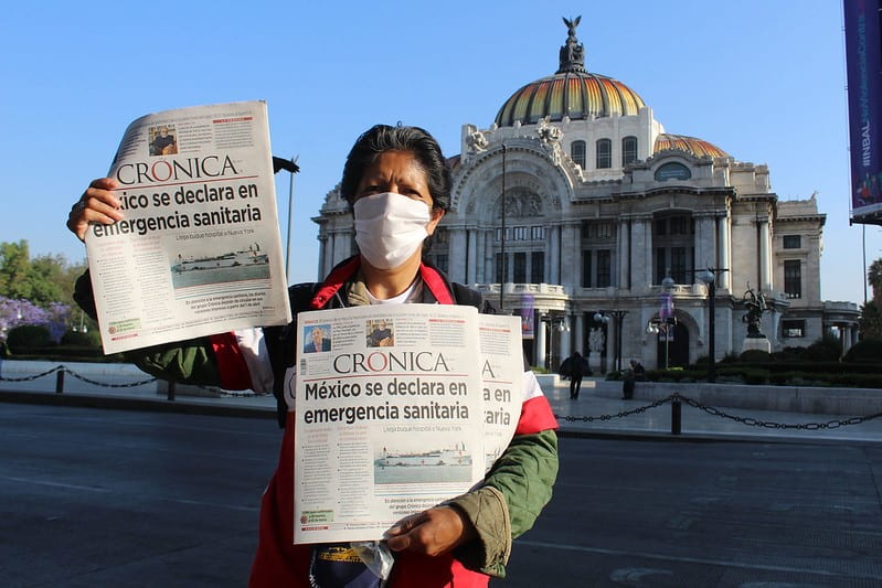 Person with a facemask holding a newspaper that says "Mexico se declara en emergencia sanitaria".