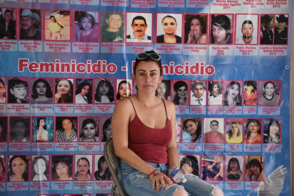 Woman sitting in front of a poster that includes pictures of femicide victims.