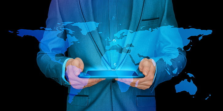 An image of a man in a blue suit holding a tablet with a hologram of the world map shining above.
