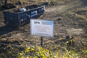 dirt field with a dumpster and a sign that reads "EPA Quanta Resources Superfund Site. Warning: Hazardous substances present in the soil. No trespassing.
