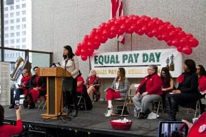 Image of woman speaking at conference with sign that reads "Equal Pay Day" in the background