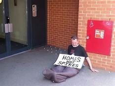 A person sitting next to a hostile architecture with a sign reading, "Homes Not Spikes"