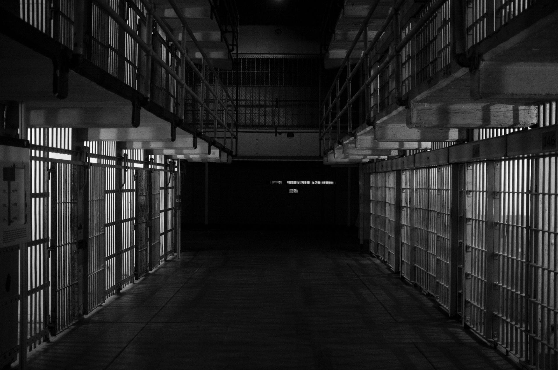 The image shows a corridor lines on either side by prison cells. There are two levels of prison cells. The prison cells have metal bars from the floor to the ceiling, and also some metal bars running along the length of the cells. Some of the entrances to the cells are open. There are no people in the image. The image has a very grim and foreboding atmosphere.