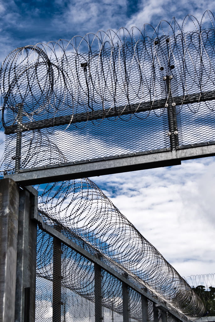 The image shows coiled, barbed wire on top of metal fences found in prisons. There is a partly cloudy sky in the background.