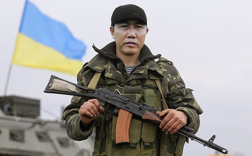 The image shows a soldier holding a gun. The Ukrainian flag is raised in the background. 