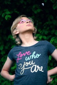 Child with a shirt that says "Love who you are." The child has their hands on their hips akin to a superhero pose and has eye makeup on.