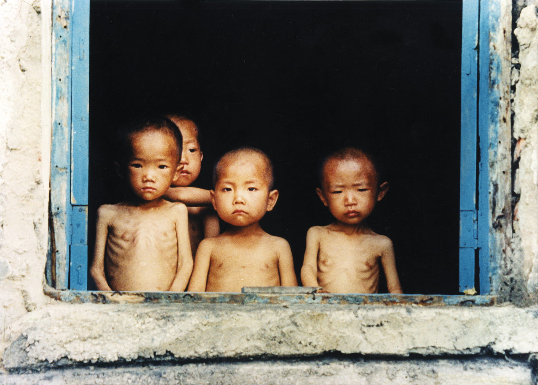 Four young Korean children stare sorrowfully through an open window with blue doors. Their ribs are visible and their arms are skinny.
