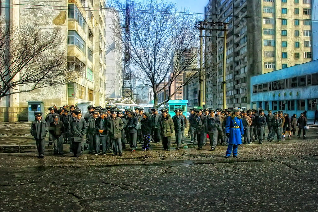 The camera is facing down a building-lined street. The buildings are neutral colors and appear old. There are two trees with no leaves. There is a group of people all wearing the same dark green/blue clothing. One person is dressed in bright blue and standing in the middle of the street.