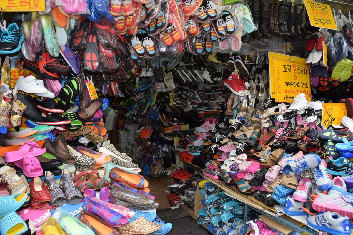 Lots of brightly colored shoes are piled onto shelves and hanging from the walls and ceilings in a Korean store. There are yellow signs with red and black text in Korean