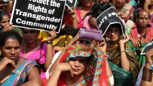 A large group of mostly women in colorful saris, jewelry, and makeup gather together with black signs with white text. One reads, "Protect the Rights of Transgender Community". 