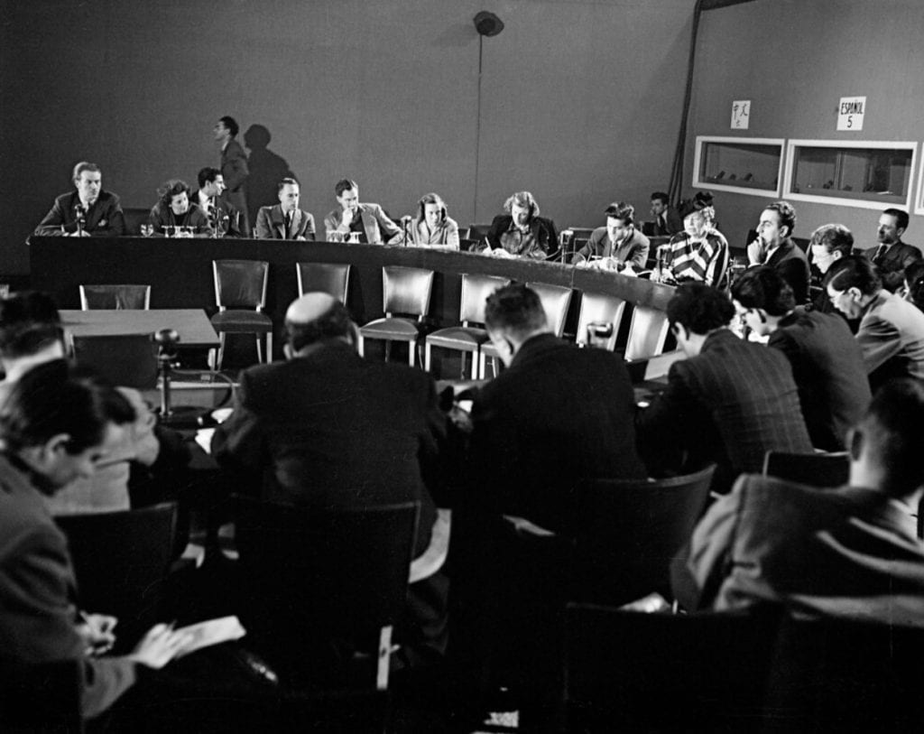 Image shows a black-and-white photograph of a press conference with people sitting around a circular table.