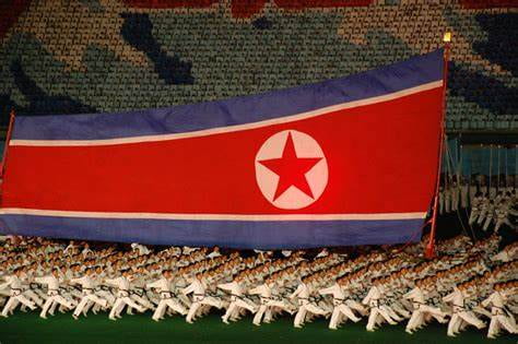 A group of people walking in sync and wearing all white uniforms. They are parading around a large North Korean flag.
