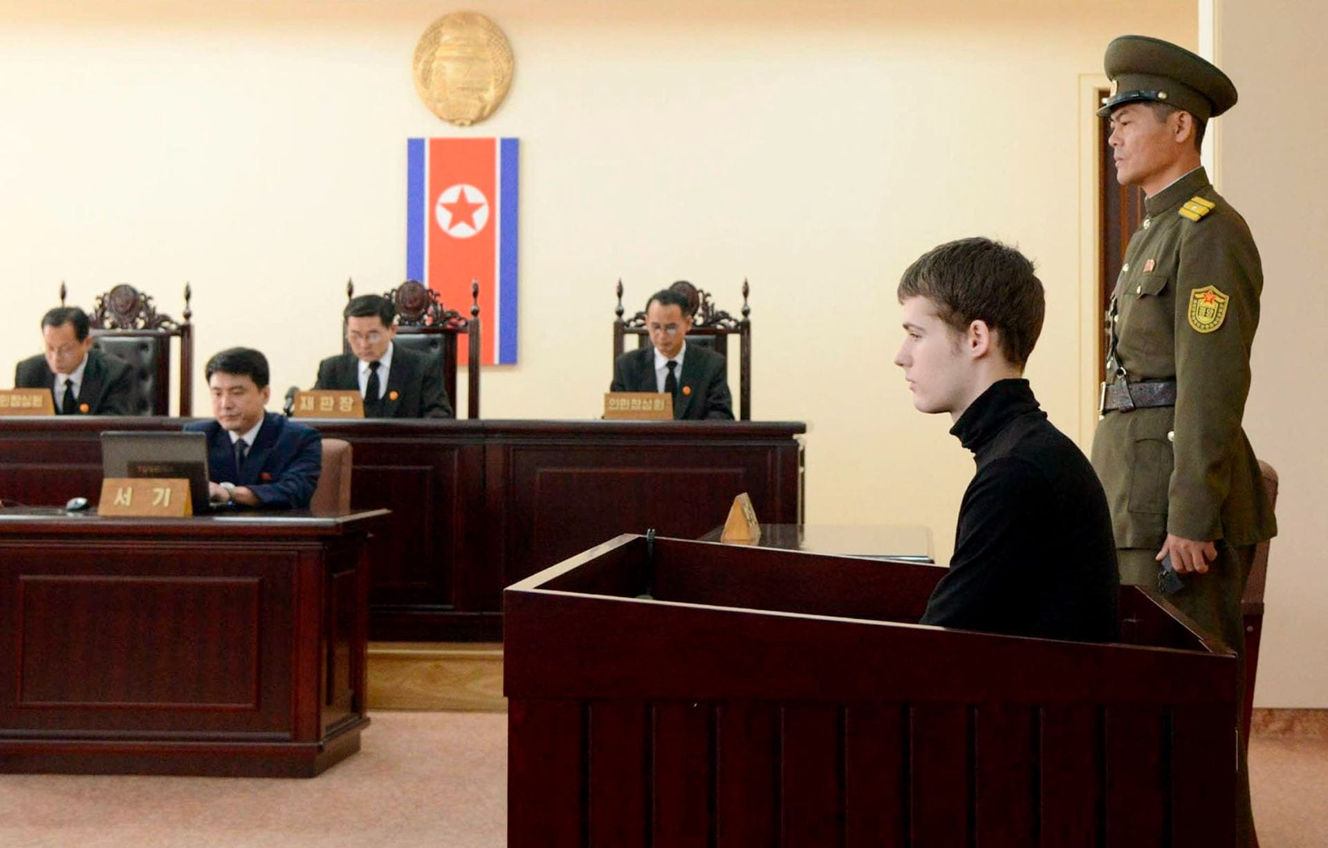 A person sits in a witness booth in front of at least four judges. There is an armed guard standing behind him, and a North Korean flag hanging on the wall behind the judges.