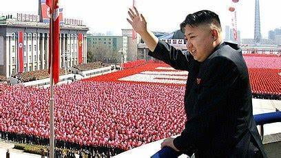Kim Jong-Un stands on a high ledge waving to thousands of people standing uniformly down below. He is wearing a black suit. Everyone in the crowd is wearing red, and there is a group of people in the distance wearing white to spell something out among the red. It is not clear what they are positioned to spell out.