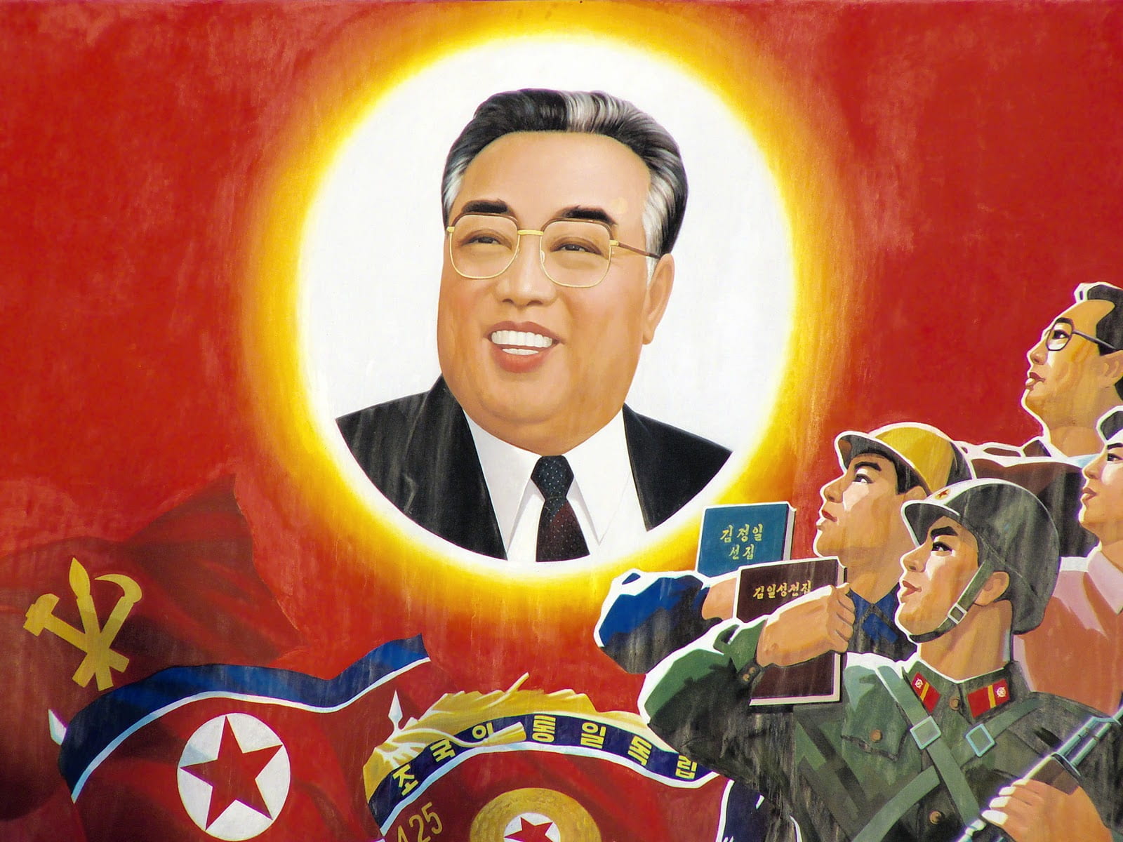 A painting with a smiling Kim Il-Sung in the center surrounded by a yellow glowing halo. In the right bottom corner, there are four Korean citizens, one of whom dressed in military attire, another in construction equipment. They are holding books and saluting the portrait of Kim Il-Sung. In the left bottom corner are three waving flags: the Workers Party of Korea, the Democratic People’s Republic of Korea, and the Korean People’s Army.