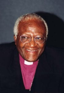 A headshot of a Black man smiling wearing purple clerical attire and collar.